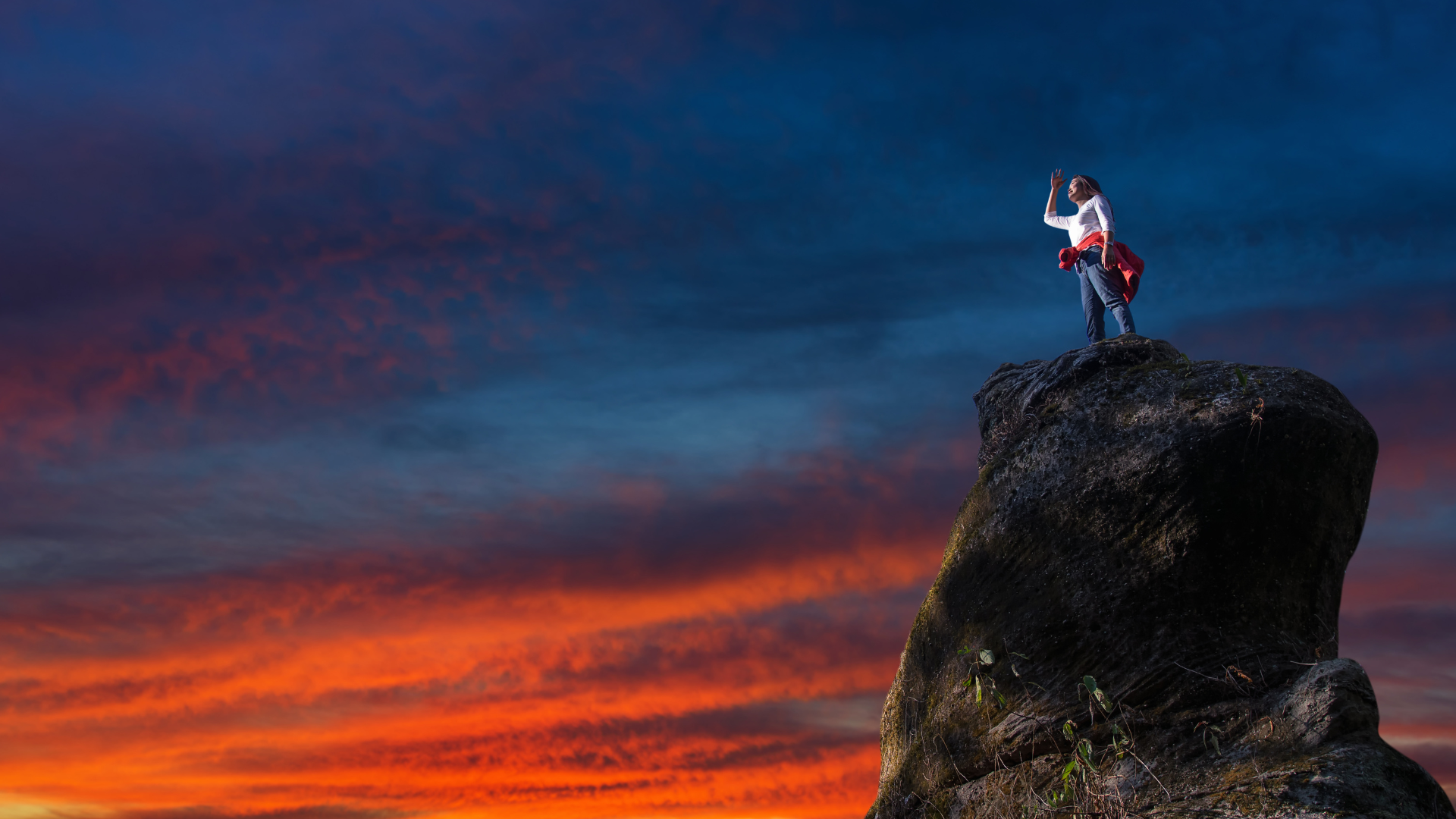 A woman stands on a large boulder and looks into the distance. In the background, the dark blue and orange colors of a sunset appear on the clouds in the sky.